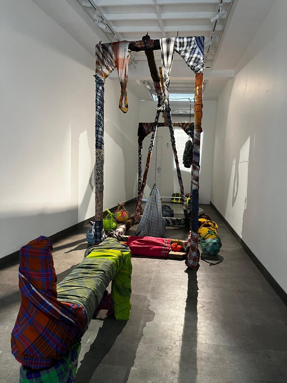 Installation view: columns and canopies of woven plaid sarongs spanning the gallery, with bundled forms resting on the floor