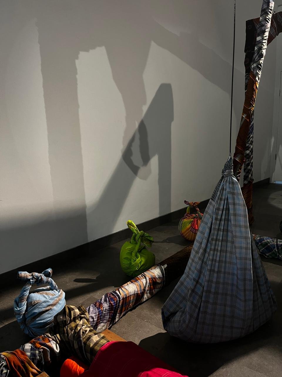 Bundled sarongs on the gallery floor, with a suspended plaid form casting a long shadow
